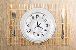 Circular clock in the form of a plate with a knife and fork next to it, with a wooden background