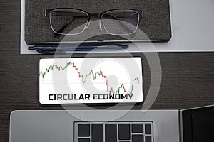 Circular economy concept. Top view of stocks price candlestick chart in phone on table near laptop, notepad and glasses with