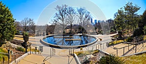 A circular stone pool with yellow winter grass, bare winter trees and lush green plants with a view of the scrapers and buildings