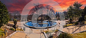 A circular stone pool with yellow winter grass, bare winter trees and lush green plants with a view of the scrapers and buildings