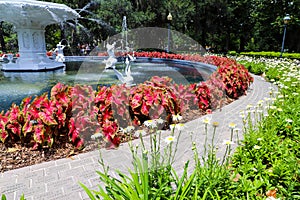 A circular white water fountain in the park surrounded by colorful flowers and lush green trees and plants with people walking