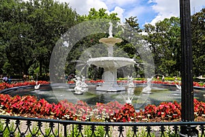 A circular white water fountain in the park surrounded by colorful flowers and lush green trees and plants with people walking