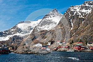 The city of Aa in Lofoten, Norway. Tiny red houses called rorbuer underneath steep snow covered mountain peaks and blue sky.