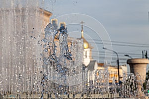 City fountain with splashes and jets of water against the background of buildings and a golden-colored church dome with a cross