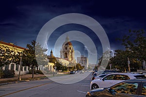 The city hall building surrounded by office buildings, lush green trees and cars on the street with storm clouds and lightning