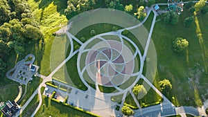 A city park covered with trees and grass, with paths for walking, in the middle of the park there is a circle paved.