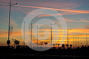 City quay with lanterns, trees and people backlit against the bright sunset sky.