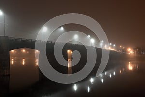 City road bridge across river in dense fog at night illuminated by lanterns