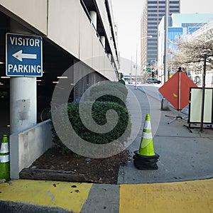 City Urbanscape and Sidewalk Scene with Visitor Parking Directional Sign, Parking Garage and Traffic Cone.