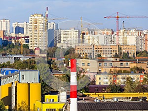 Cityscape with cranes, high-rise buildings, construction sites and factory pipe