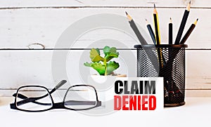 CLAIM DENIED written on a white business card next to pencils in a stand and glasses. Nearby is a potted plant