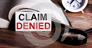 CLAIM DENIED written on a white card near an open book, alarm clock and magnifying glass