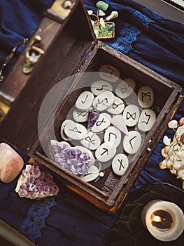 Clairvoyant tools rune stones, crystal pendulums in natural dark wooden case box on dark blue background.