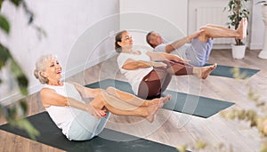 Class of elderly sporty women stretching gluteal muscle while lying on back during yoga Pilates workout on mats in gym