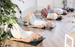 Class of elderly sporty women stretching gluteal muscle while lying on back during yoga Pilates workout on mats in gym