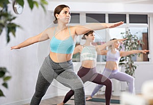 Class of focused women standing in Virabhadrasana 2 or warrior 2 position during group yoga training in gym studio