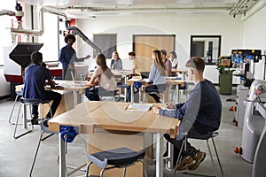 Class Of High School Students Sitting At Work Benches Listening To Teacher In Design And Technology Lesson