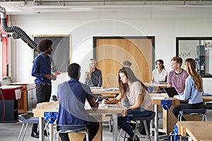 Class Of High School Students Sitting At Work Benches Listening To Teacher In Design And Technology Lesson