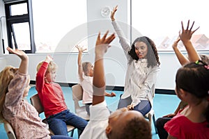 A class of infant school children sitting on chairs in a circle in the classroom, raising hands with their female teacher, close u