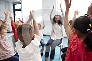 A class of infant school children sitting on chairs in a circle in the classroom, raising hands with their female teacher, close u