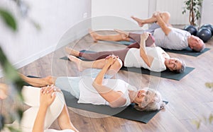 Class of mature women stretching gluteal muscle while lying on back during yoga Pilates workout on mats in gym studio
