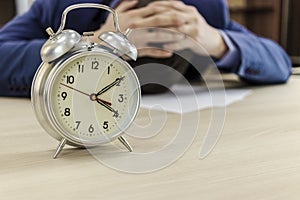 Classic alarm clock on the office table. In the background, the manager laid his head on the table and covered it with his hands.