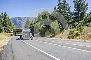 Classic American big rig semi truck with chrome parts and empty flat bed semi trailer driving on the winding road with arched