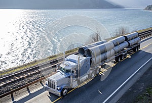 Classic American big rig semi truck transporting big plastic pipes on flat bed semi trailer driving on the road along the river in