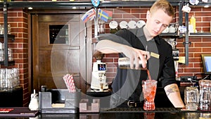 Classic bartender in black uniform pouring colorful liquid from shaker to a cocktail glass in interior classy bar
