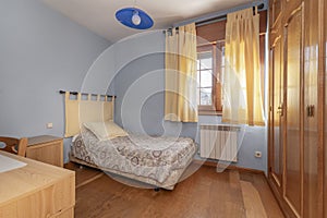 Classic bedroom with a single bed covered with a flower-print bedspread, a wooden window with curtains and light blue painted