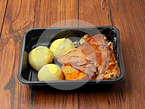 Classic beef dinner meal with slice of meat, mashed potatoes, gravy and turnip in dark plastic container on wooden table