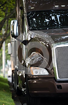 Classic big rig bonnet dark brown semi truck tractor standing in front of another semi trucks on the road side with green trees