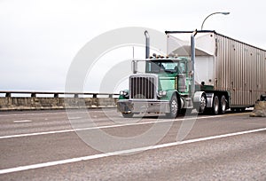 Classic big rig day cab semi truck with corrugated bulk semi trailer driving on the wide highway