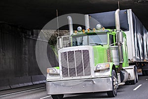 Classic big rig green semi truck transporting commercial cargo in covered black semi trailer running under the bridge across wide