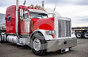 Classic big rig red semi truck tractor with chrome accessories and flat bed semi trailer standing on parking lot waiting for load