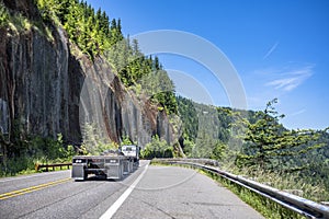 Classic big rig semi truck with empty flat bed semi tailer running to warehouse on the winding mountain road with rock cliff and