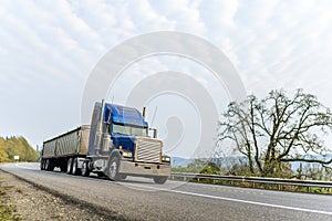 Classic blue big rig American bonnet semi truck driving with covered bulk semi trailer on the winding autumn road