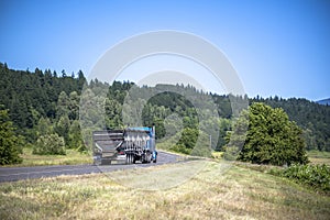 Classic blue big rig semi truck with covered bulk semi trailer running on the narrow winding road with summer meadows and hills