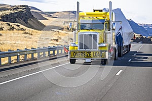 Classic bright yellow powerful big rig semi truck with oversize load sign on the bumper transporting oversized cargo on step down