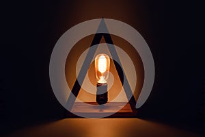 Classic Edison old glowing light bulb with filament in loft lamp standing in wooden triangl as a candle. Abstract