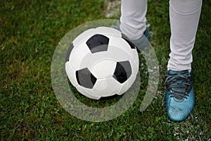 A classic football ball and feet of a young boy on a field background. Children training soccer. A ball on a grass.
