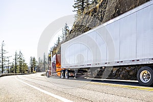 Classic orange big rig semi-truck tractor transporting cargo in dry van semi trailer driving on the mountain highway road with