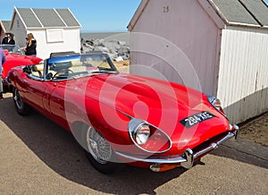 Classic Red E Type Jaguar Open Top Sports car parked on seafront promenade near beach huts.