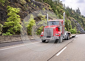 Classic red industrial big rig day cab semi truck tractor transporting empty step down semi trailer climbing on the mountain road