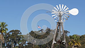 Classic retro windmill, bladed rotor and USA flag against blue sky. Vintage water pump wind turbine, power generator on livestock