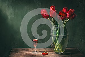 Classic still life with beautiful red tulip flowers bouquet in transparent glass vase and a glass of red wine. Art photography