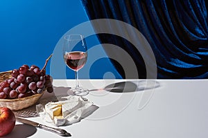 Classic still life with fruits, red wine and Camembert cheese on white table near velour curtain isolated on blue