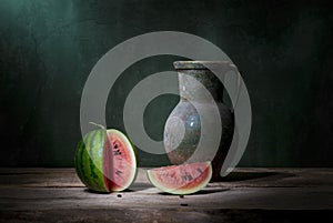 Classic still life with one small green cut watermelon and a jug. Art photography.