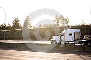 Classic style big rig white semi truck tractor with flat bed semi trailer running on the sunny divided highway road