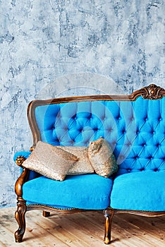 Classic vintage blue buttoned sofa with golden cushions on gray concrete wall indoors, copy space. Living room interior.